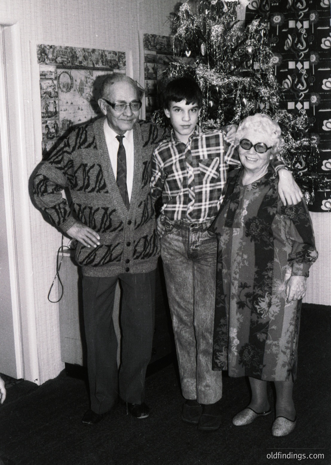 Family portrait from the **1970s**, featuring a man in a patterned sweater and tie, a young boy in plaid shirt and bell-bottoms, and a woman in a floral dress with sunglasses. Decor includes a Christmas tree with geometric ornaments and framed artwork. Warm, intimate home setting.