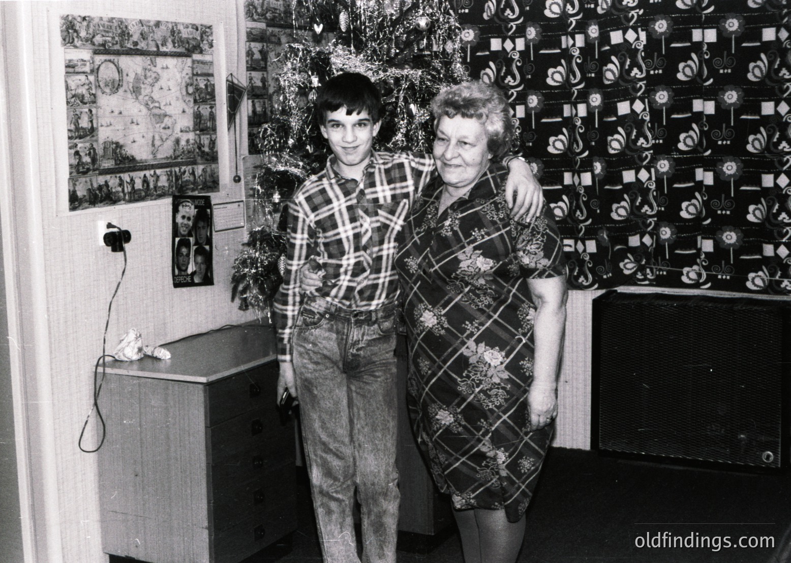 A black-and-white family portrait from the 1970s–1980s, featuring a young man and woman in patterned clothing standing indoors. The man wears a checkered shirt and baggy pants; the woman sports a floral-patterned dress. Decor includes wallpaper with geometric designs, a framed map/poster, and a small wooden cabinet. Warm, intimate home setting reflects mid-century domestic life.