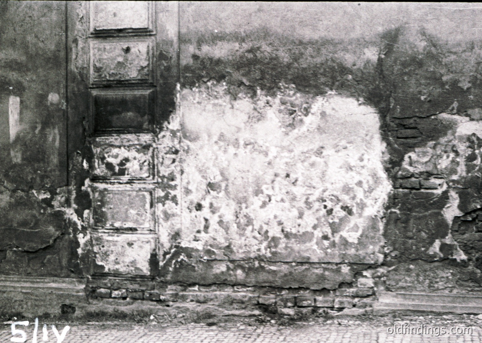 War-damaged brick facade with plaster collapse, exposing rough stonework. Likely Eastern European urban setting, mid-20th century. Evidence of shelling or bombing.
