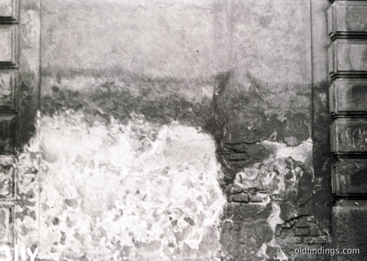 Vintage sepia-toned photograph showing a collapsed stone wall and debris, likely from an earthquake or structural failure. The scene captures partial ruins with exposed bricks and rubble, framed by intact architectural columns on either side. The hazy background suggests distant structures or terrain.
