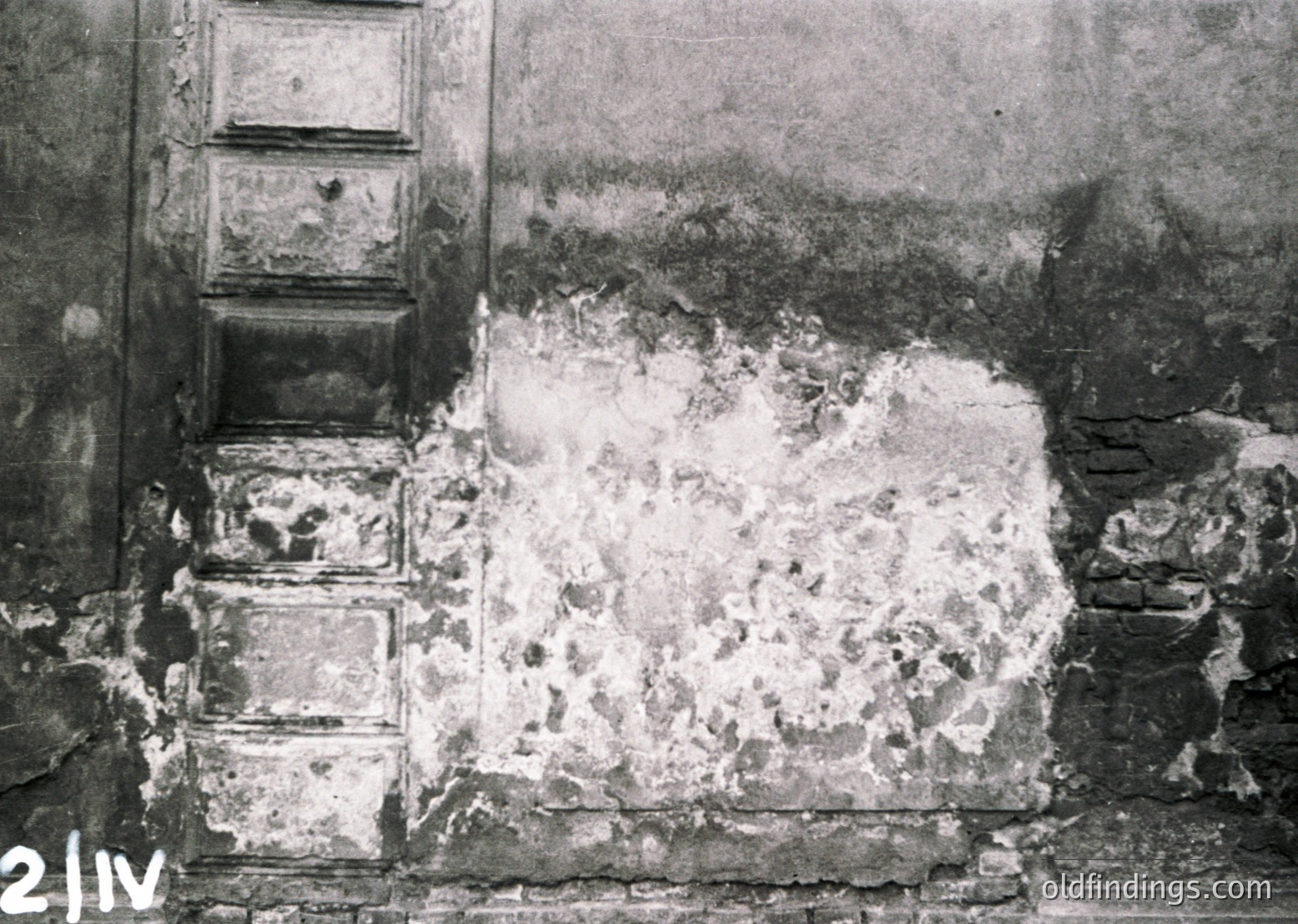 Black-and-white close-up of a damaged plaster wall with exposed brickwork and crumbling debris, likely from a bomb or artillery strike. Vertical wooden paneling remains intact on the left. Mid-20th century wartime architecture.