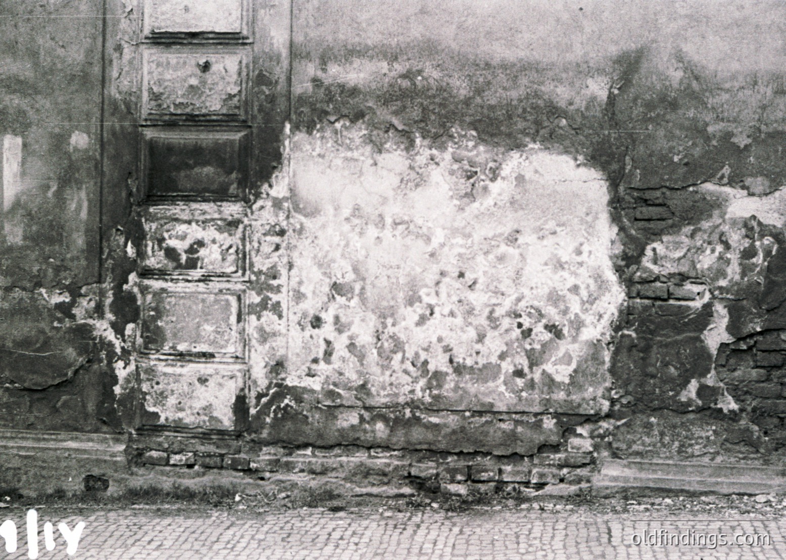 War-damaged brick facade with exposed plaster and mortar, likely post-WWII. Cobblestone street and partial stone archway visible. Architectural detail suggests European urban setting, possibly 1940s–1950s.
