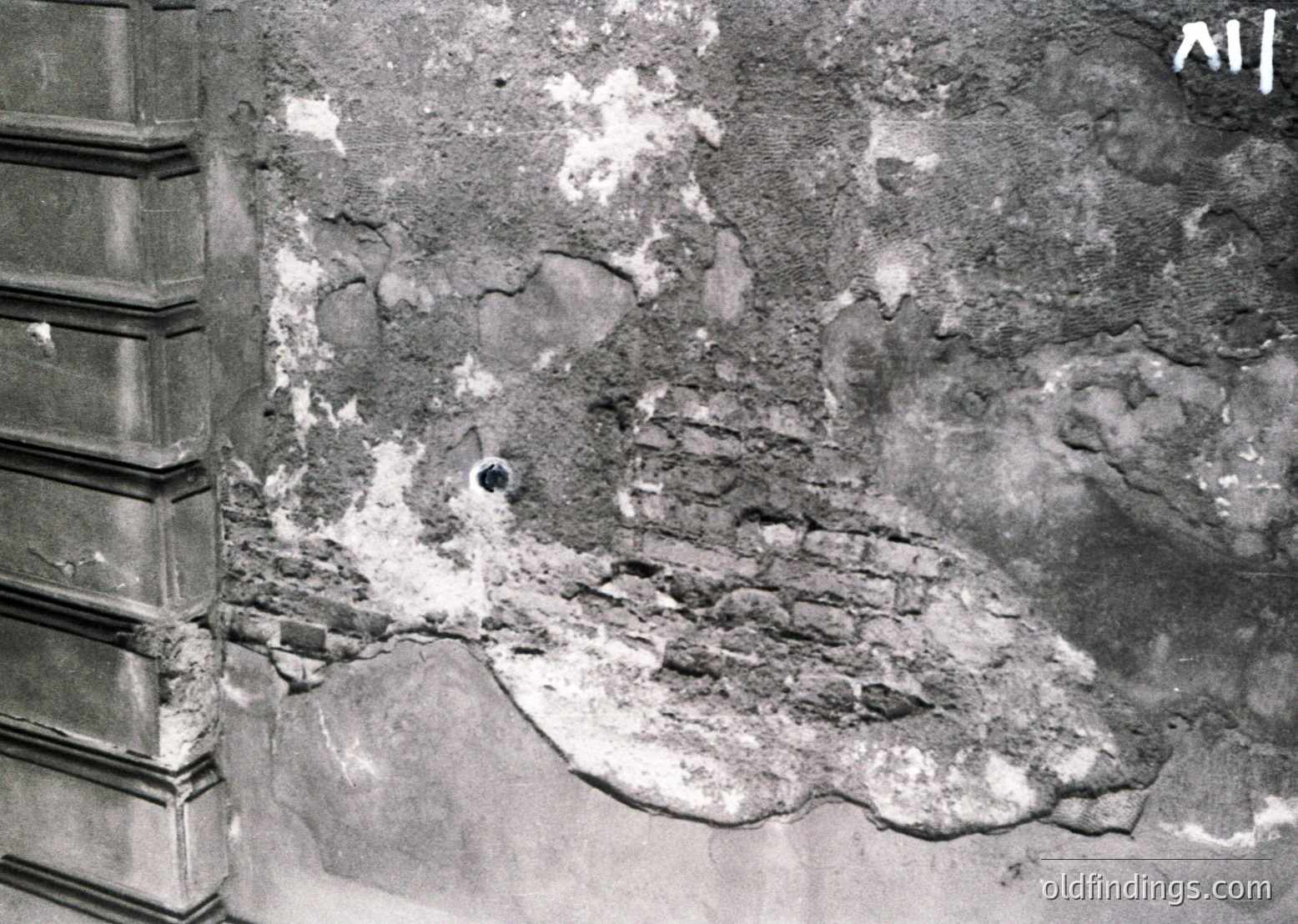 Close-up of severely damaged plaster and exposed brickwork, likely from a bomb or artillery strike. The jagged, uneven surface reveals structural compromise, with debris embedded in the wall. Architectural detail includes a partial column base on the left.