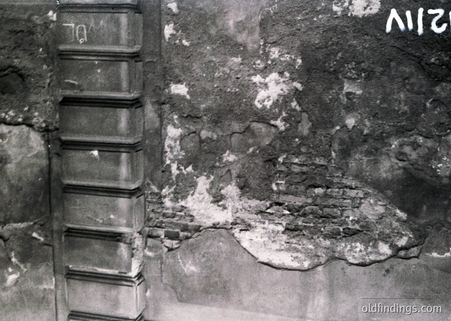 Black-and-white close-up of weathered concrete wall showing severe structural damage—exposed rebar and cracked plaster. Likely post-war or industrial ruin, indicative of mid-20th century construction neglect.