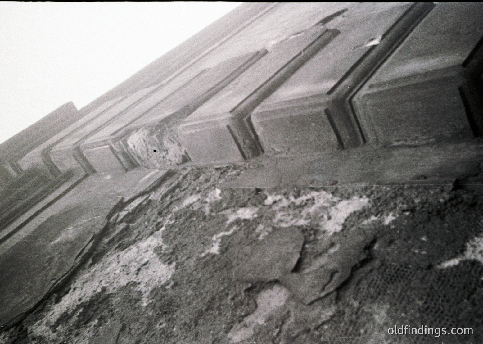 Close-up of weathered concrete ledge with exposed rebar and cracked surface, likely part of an industrial or urban structure. The texture and erosion suggest prolonged exposure to elements, possibly mid-20th century construction.