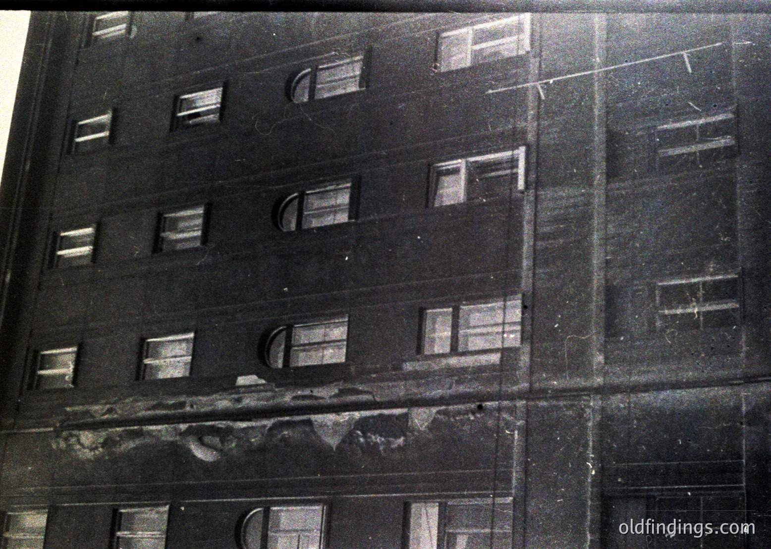 Multi-story brick apartment building facade, likely mid-20th century, showing signs of wear—cracks, exposed mortar, and peeling plaster. Symmetrical arched windows with metal grilles, indicative of Soviet-era construction. Urban decay evident in weathered exterior.