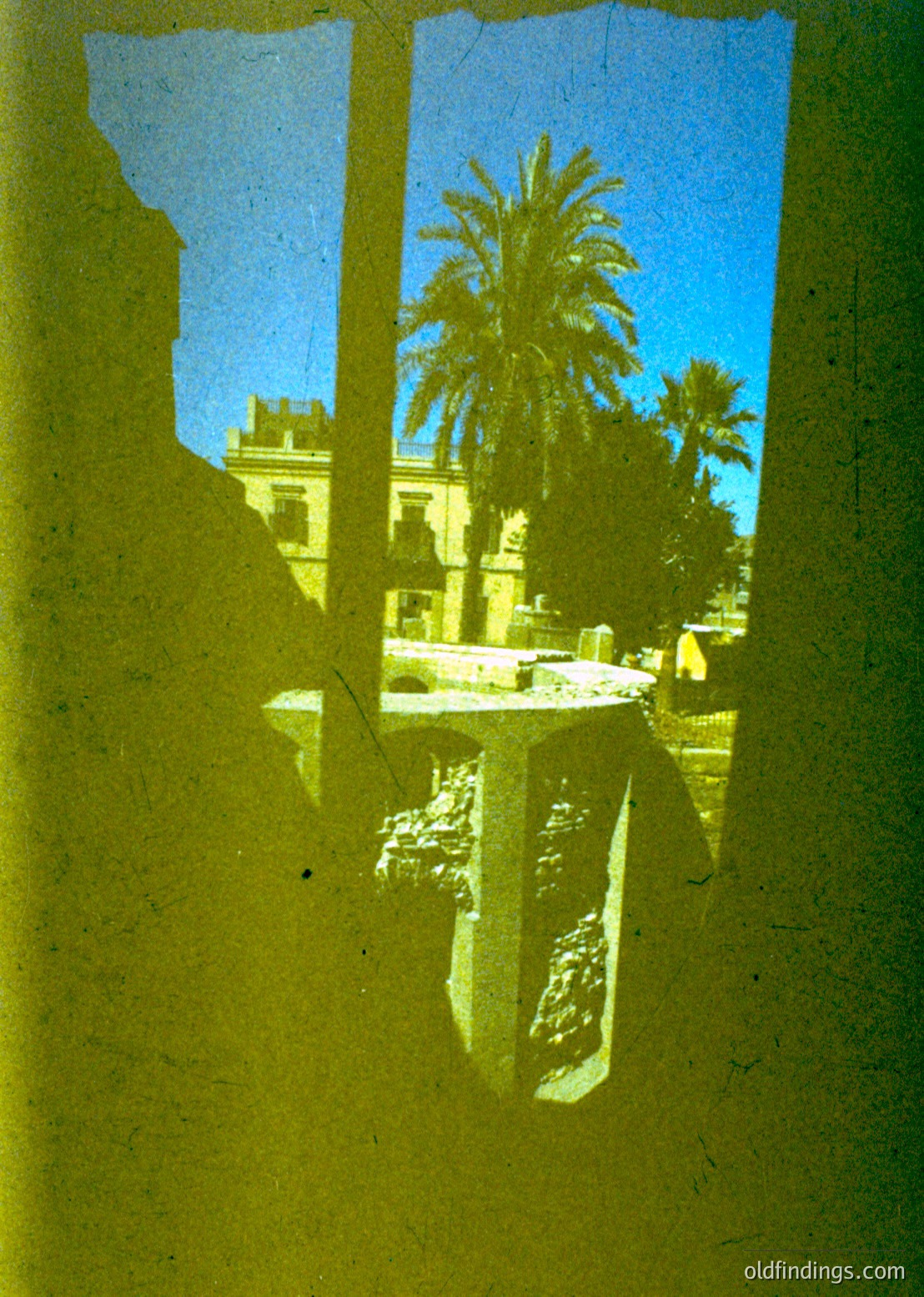 Vintage sepia-toned view through a window frame revealing a Mediterranean courtyard. Palm trees, classical stone columns, and a distant building suggest a historic or colonial-era residence. Yellowed paper texture indicates an aged photograph, likely from the early-to-mid 20th century.