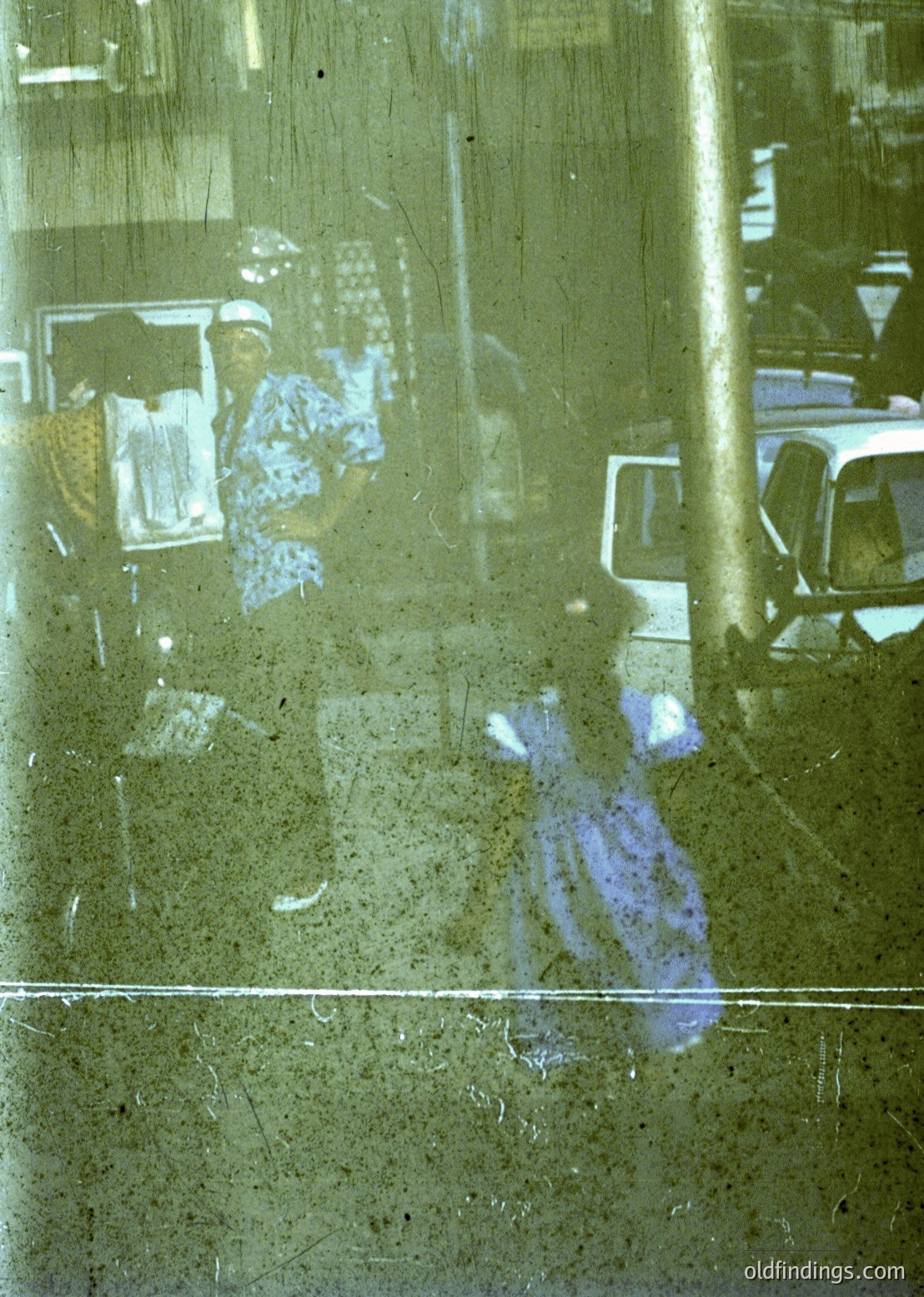 Reflection of a 1970s-era bus station interior: blurred passengers seated on patterned upholstery, a white van parked outside, and a blue garment silhouette on a wet floor. Yellowed glass and aged lighting fixtures suggest institutional architecture.