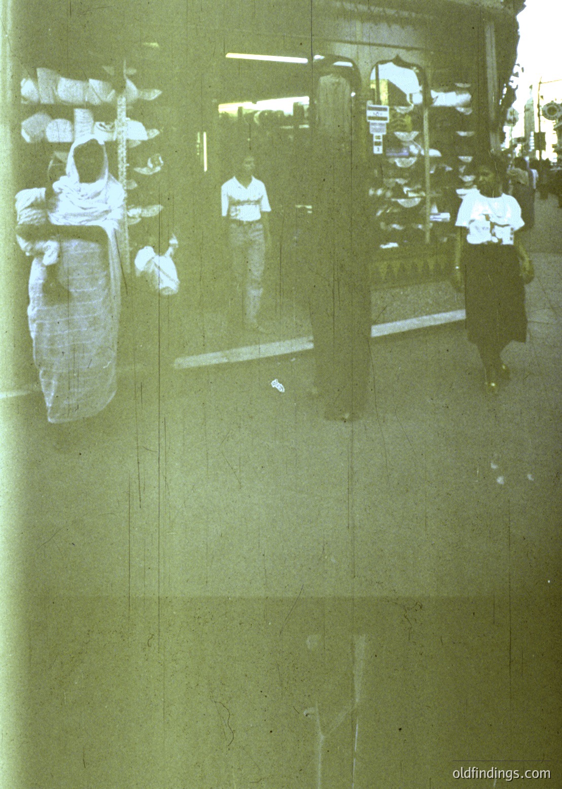 Reflection of 1970s urban street scene in wet pavement, showing blurred figures in casual attire (white shirts, dark pants) and a hanging laundry rack with folded clothes. Industrial architecture with metal railings and storefronts visible.