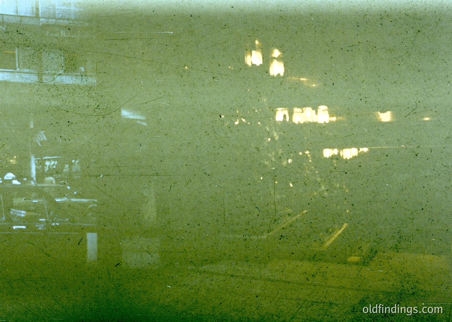 Vintage close-up of a foggy, rain-streaked window reflecting blurred city lights and indistinct architecture. Warm streetlights and distant buildings create a hazy, nostalgic urban scene. Likely mid-20th century, possibly or .
