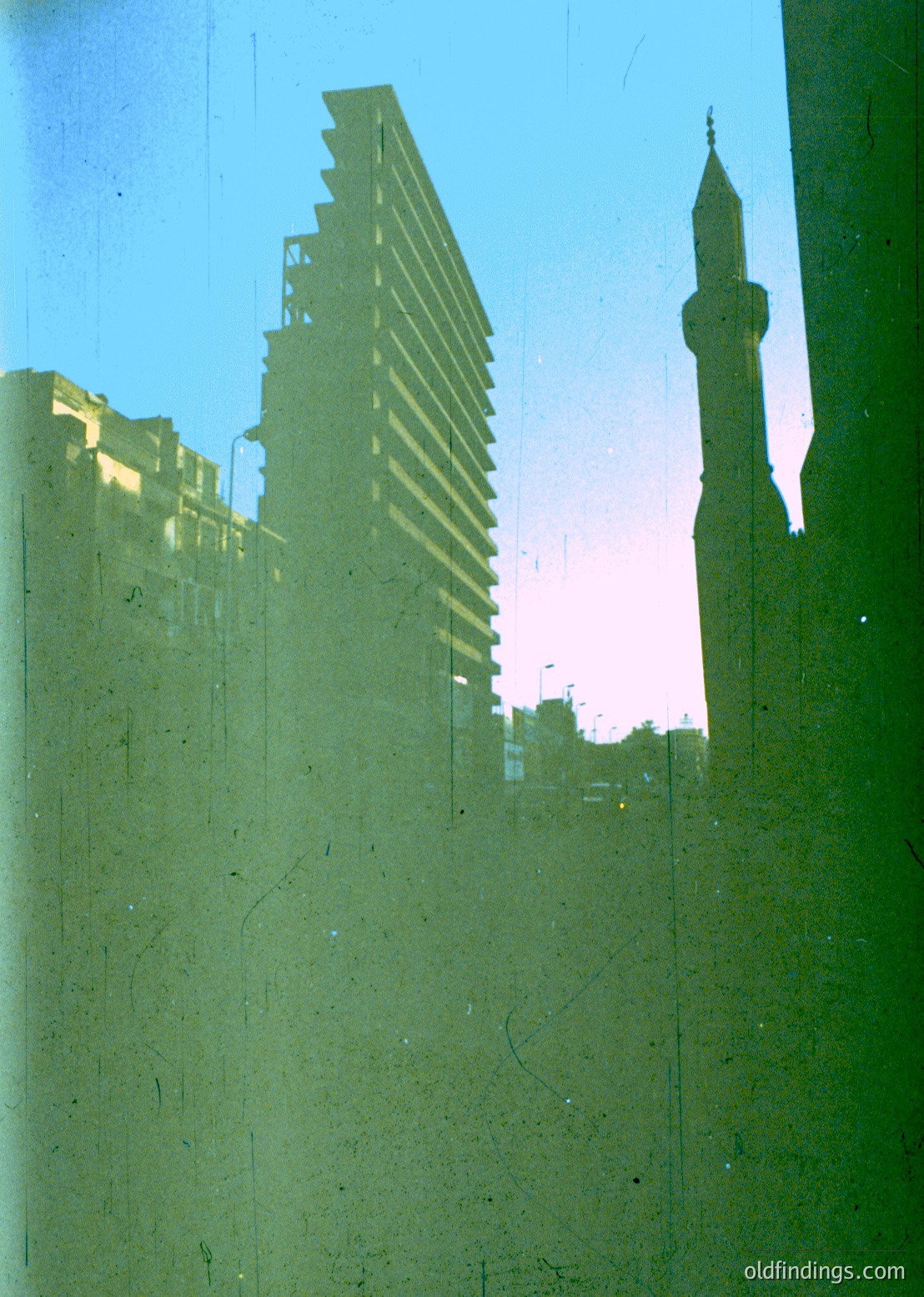 Mid-century modernist high-rise framed by Islamic minaret, likely –70s urban architecture. Concrete Brutalist facade contrasts with slender, pointed minaret silhouette. Reflective glass or film grain suggests archival or vintage photography. Potential or setting.
