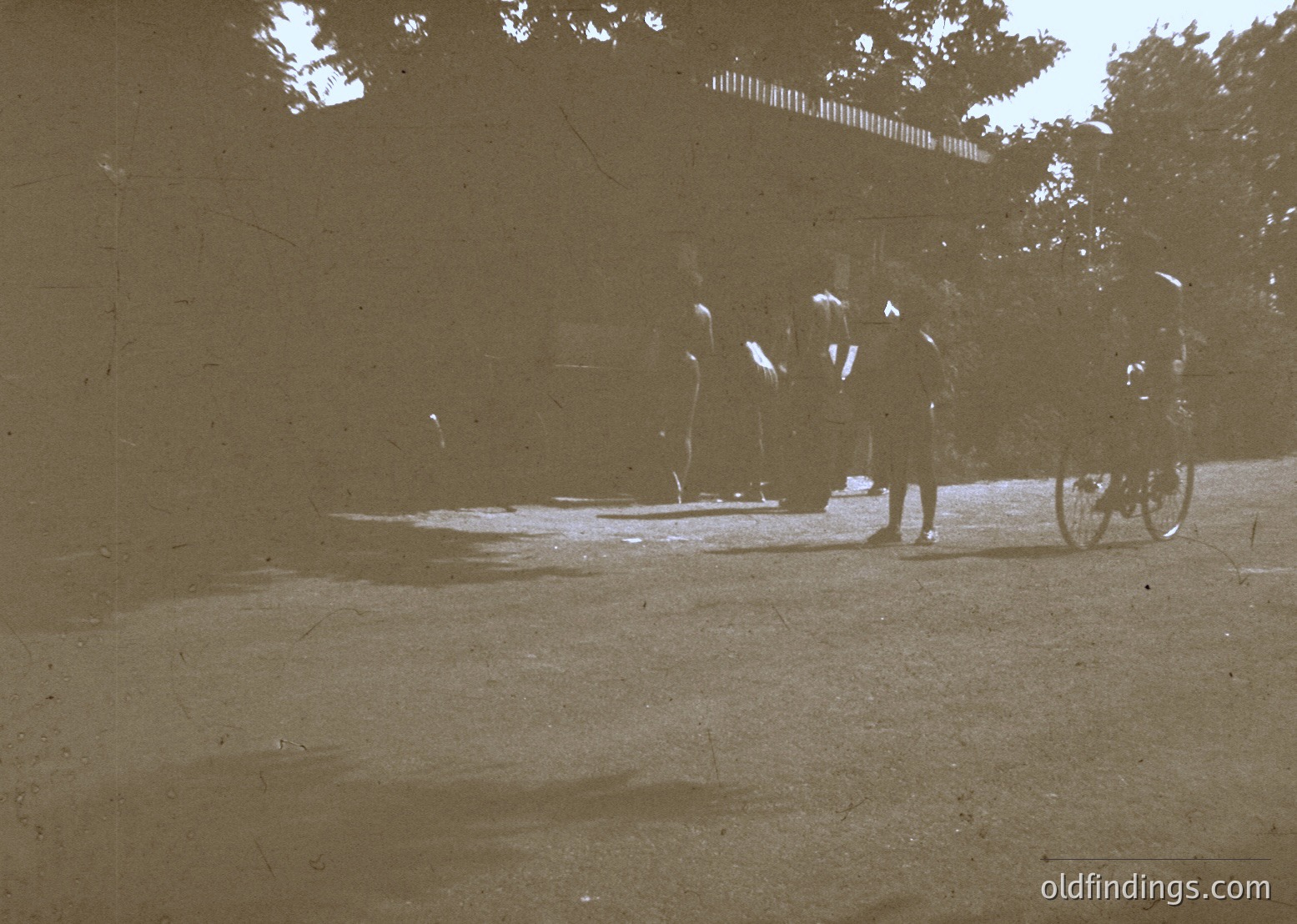 Vintage sepia-toned street scene showing three figures in early 20th-century attire—long coats, hats, and bicycles—walking near a tree-lined sidewalk. Blurred signage hints at a European urban setting.
