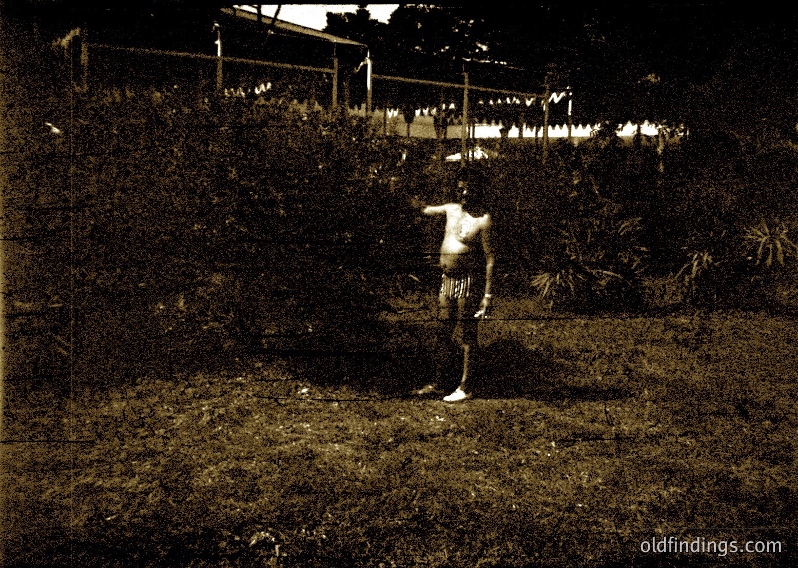 Vintage sepia-toned photograph of a shirtless man in motion, likely mid-throw, in a lush garden setting. Clothesline with hanging laundry in background suggests domestic activity. Style and lighting suggest early 20th-century amateur photography.