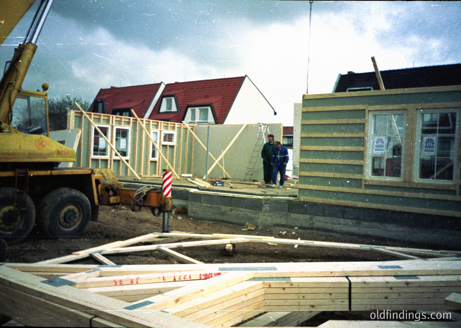 Mid-construction residential site featuring skeletal timber-frame homes ( ). Two workers in workwear inspect framing, with a yellow crane truck in foreground. Partial view of finished roof and windows suggests 1980s-1990s suburban development ( ). Ground-level lumber and concrete footings indicate early-stage construction.