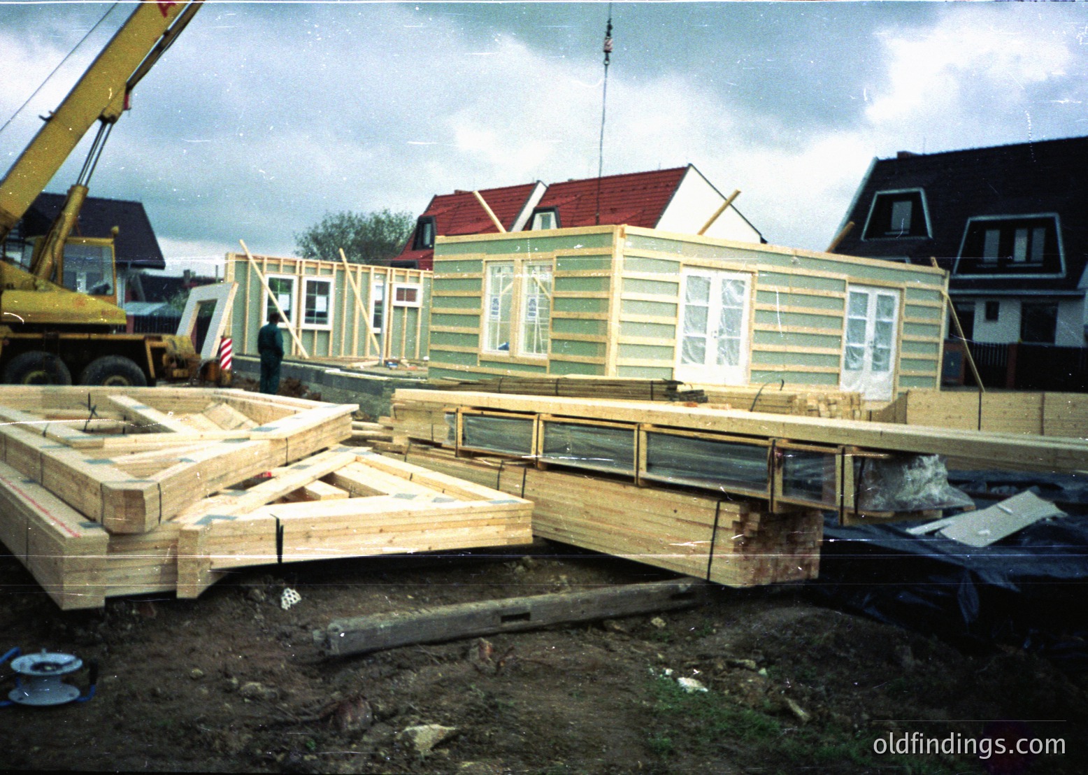 Modular prefab home under construction, elevated on wooden supports with crane assistance. Light green siding and large windows visible. Surrounding residential neighborhood with pitched roofs. Likely mid-20th century construction method.