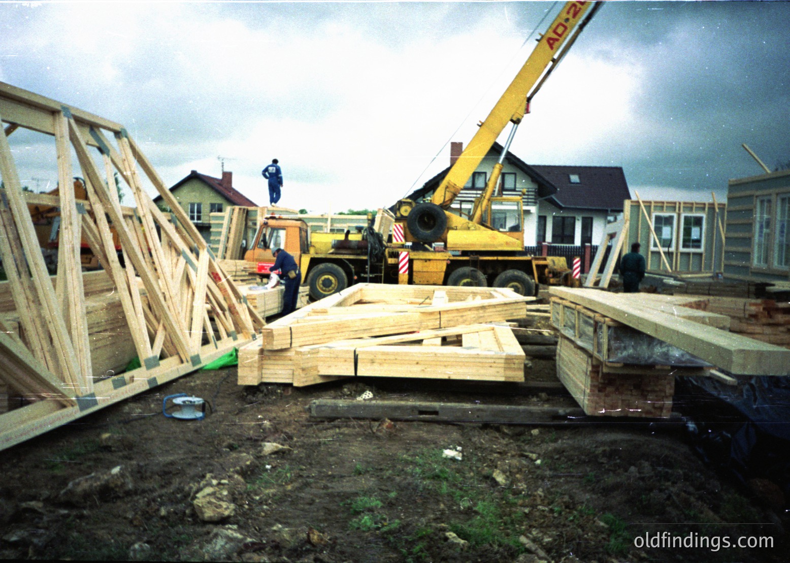 Construction site featuring a yellow ADHER crane lifting prefabricated wooden beams. Workers in blue uniforms oversee the assembly of modular housing units. Residential structures in background suggest suburban development. Likely late 20th century (1980s-1990s) based on equipment and attire.