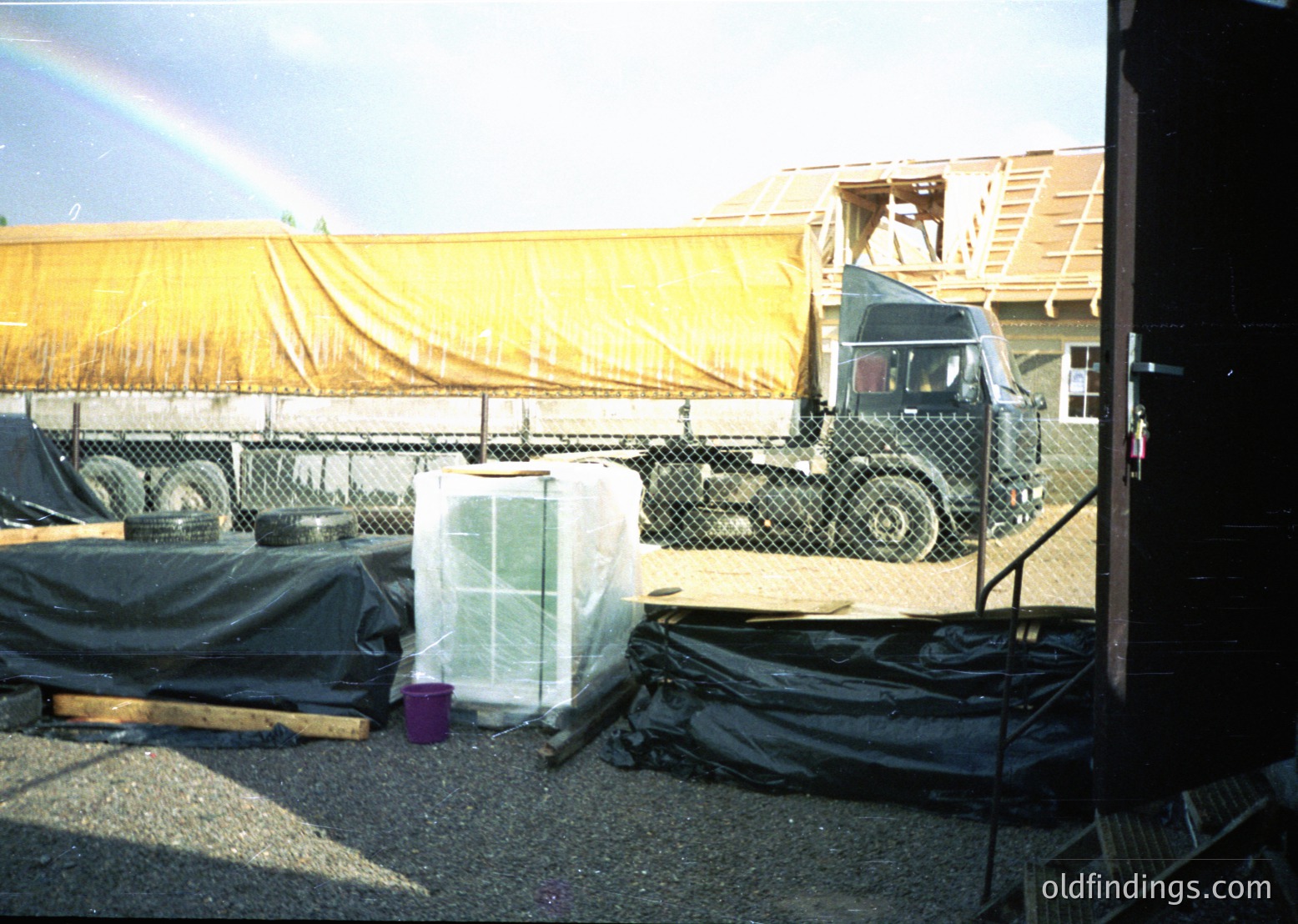 Industrial site featuring a military-style truck with a covered cargo area, likely for transporting heavy or secured materials. A large transparent plastic-wrapped object sits on a pallet beside the truck. Tarps and black plastic sheeting cover other materials, suggesting logistics or construction activity. Fencing and a partial building frame indicate a temporary or semi-permanent setup. Sunlight creates a rainbow in the background, adding a striking contrast to the utilitarian scene.