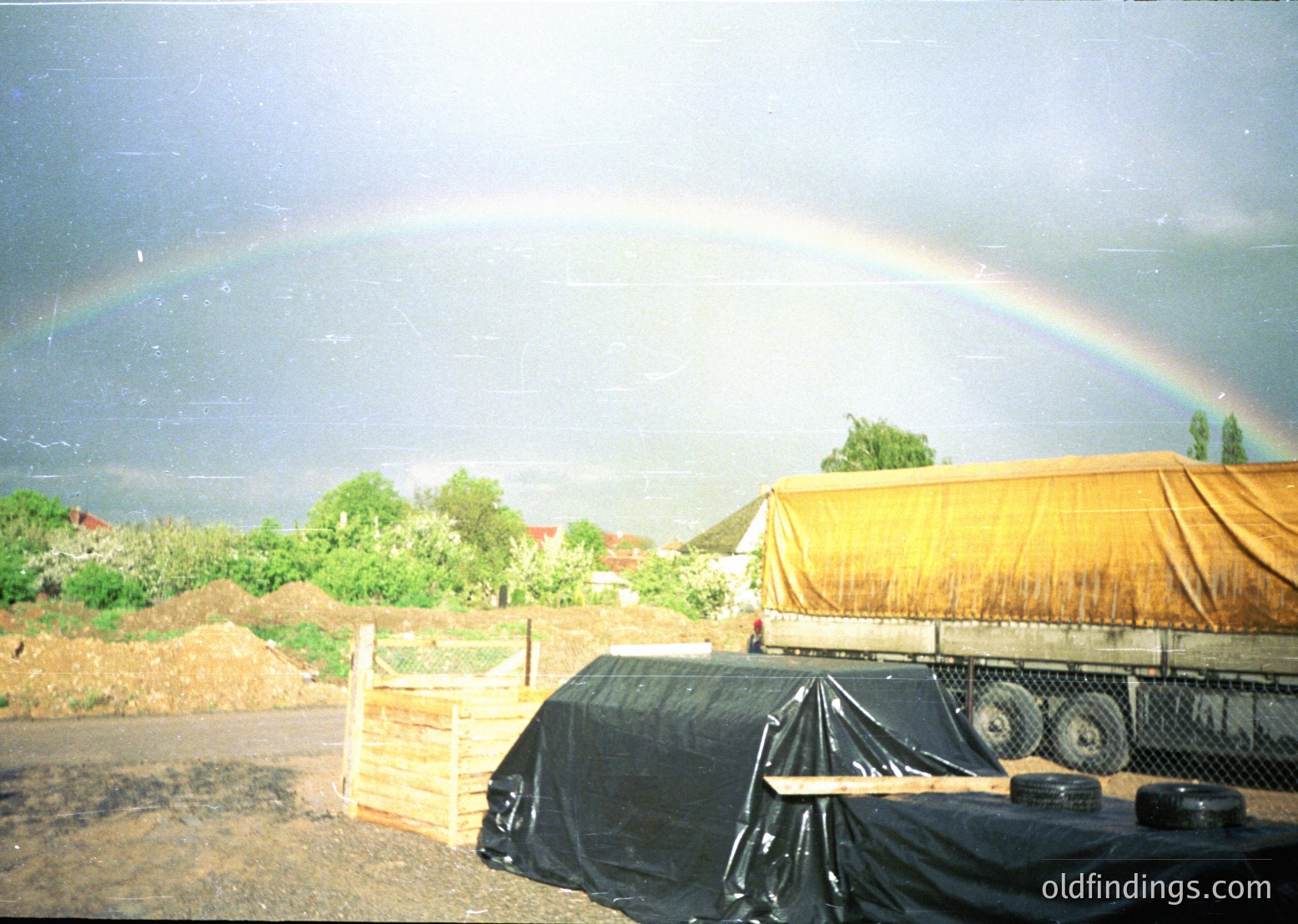 Double rainbow arcs over a rural construction site with tarpaulin-covered trucks and dirt piles.