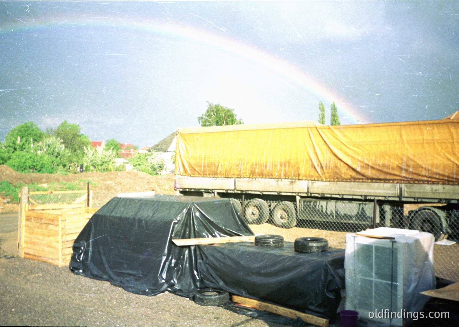 A vintage truck with a yellow tarp covers its cargo area, parked beside a wooden fence. Black plastic sheeting drapes over stacked tires and crates. A vivid rainbow arcs over a rural landscape with scattered buildings and greenery. Likely late 20th century, possibly Eastern Europe.