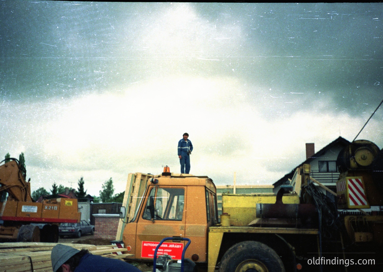 Construction worker stands atop yellow JCB excavator in industrial yard, 1970s-80s. Overcast skies and heavy machinery—including a red-and-white striped trailer—suggest mid-century European site. Ideal for industrial heritage, machinery archives, or historical construction documentation.