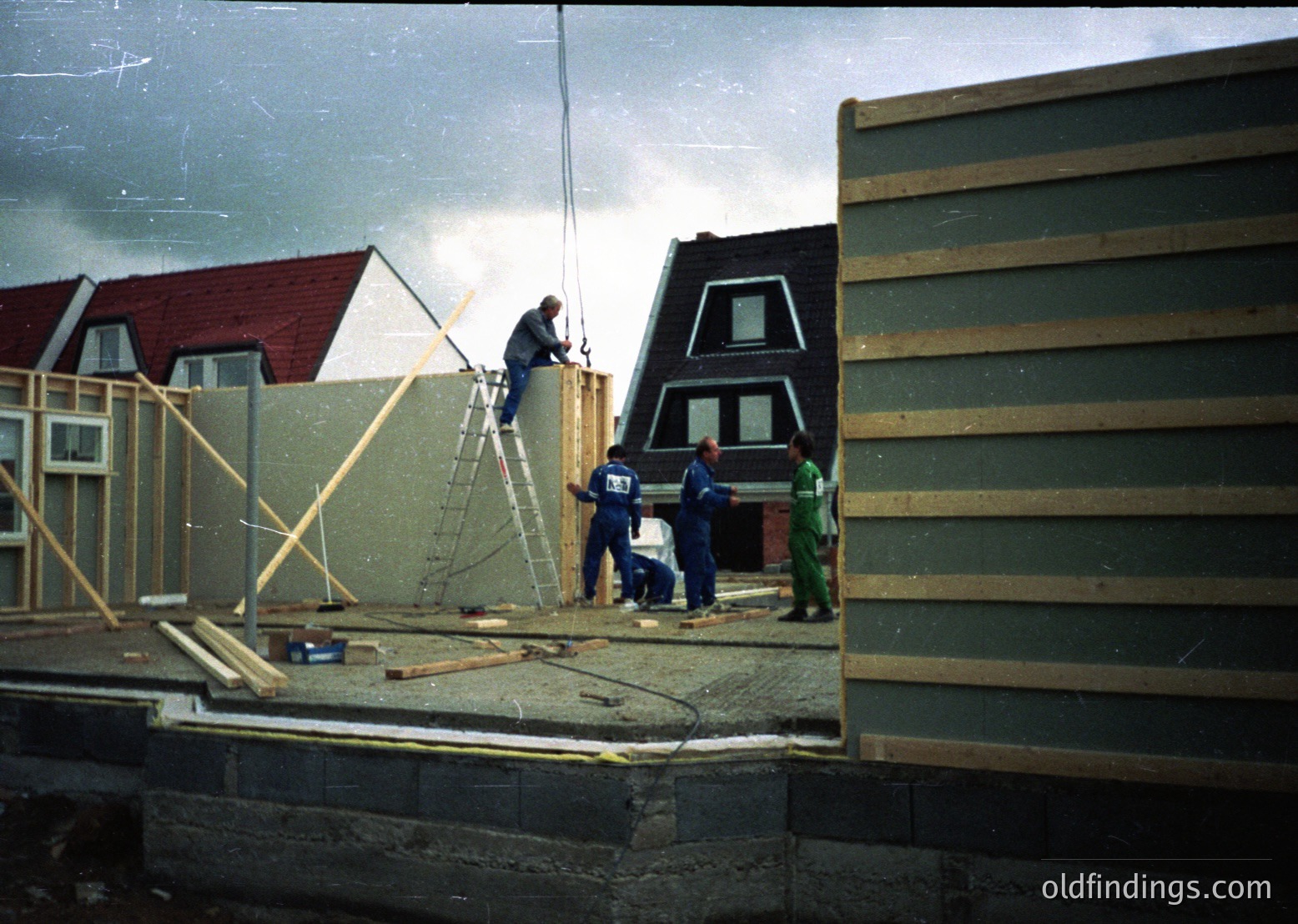 Construction crew erects prefab panel walls on a residential site, likely mid-20th century. Workers in overalls and hard hats use scaffolding and ladders to assemble modular housing units. Adjacent homes feature gabled roofs and white-framed windows, suggesting suburban development. Vintage color tone and grain indicate possible or era.