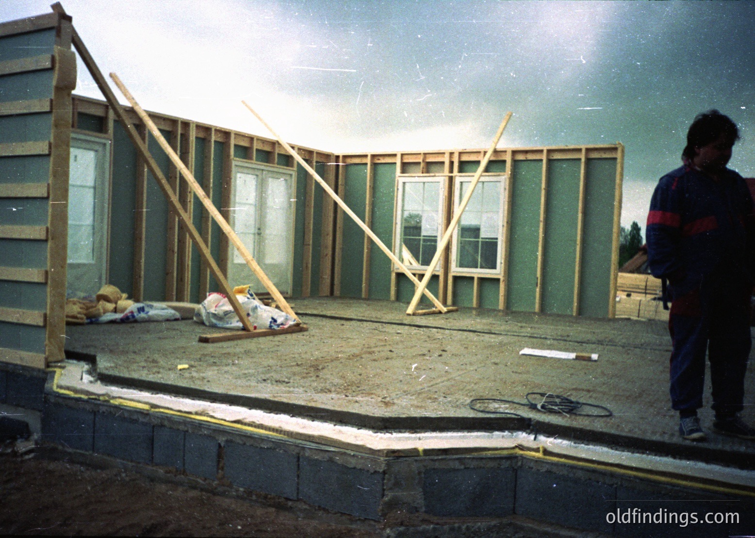 Mid-stage construction of a single-story home, featuring green insulated panels and wooden framing. A worker in dark pants and a striped shirt stands near the foundation, which includes poured concrete and brickwork. Sunlight illuminates the scene, casting shadows on the partially framed walls and windows.