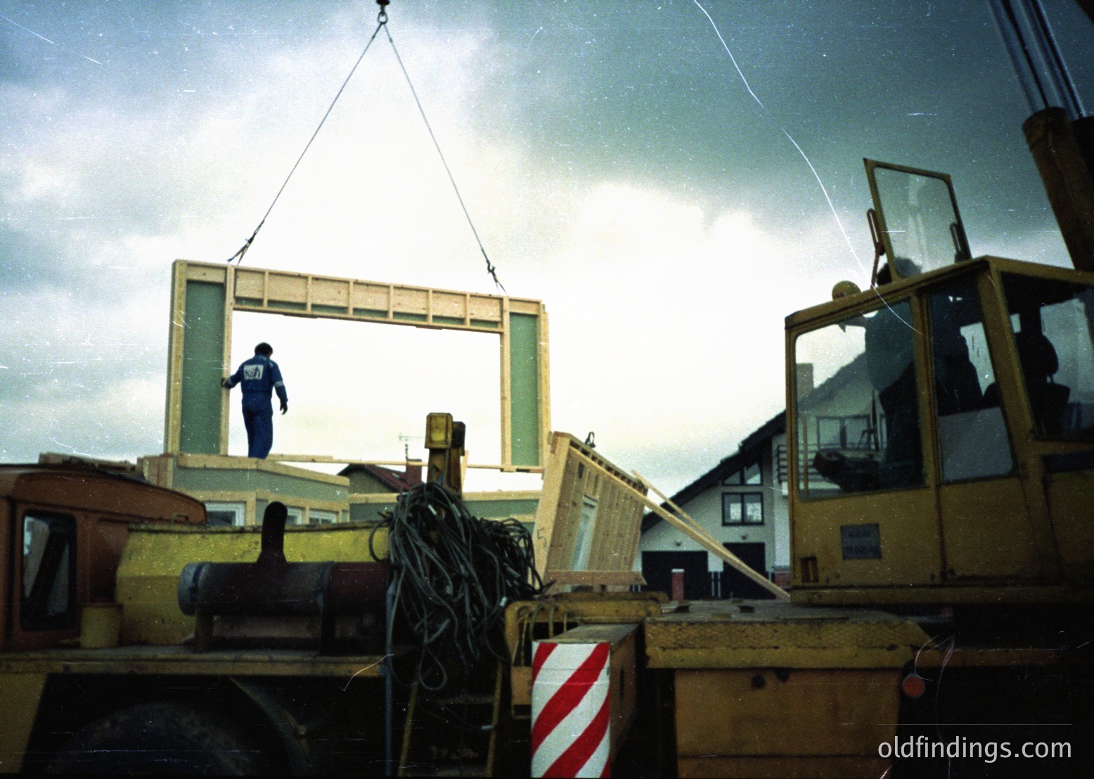 Construction worker hoists large wooden window frame via crane at industrial site, likely mid-20th century. Overcast skies and heavy equipment suggest heavy lifting operations.