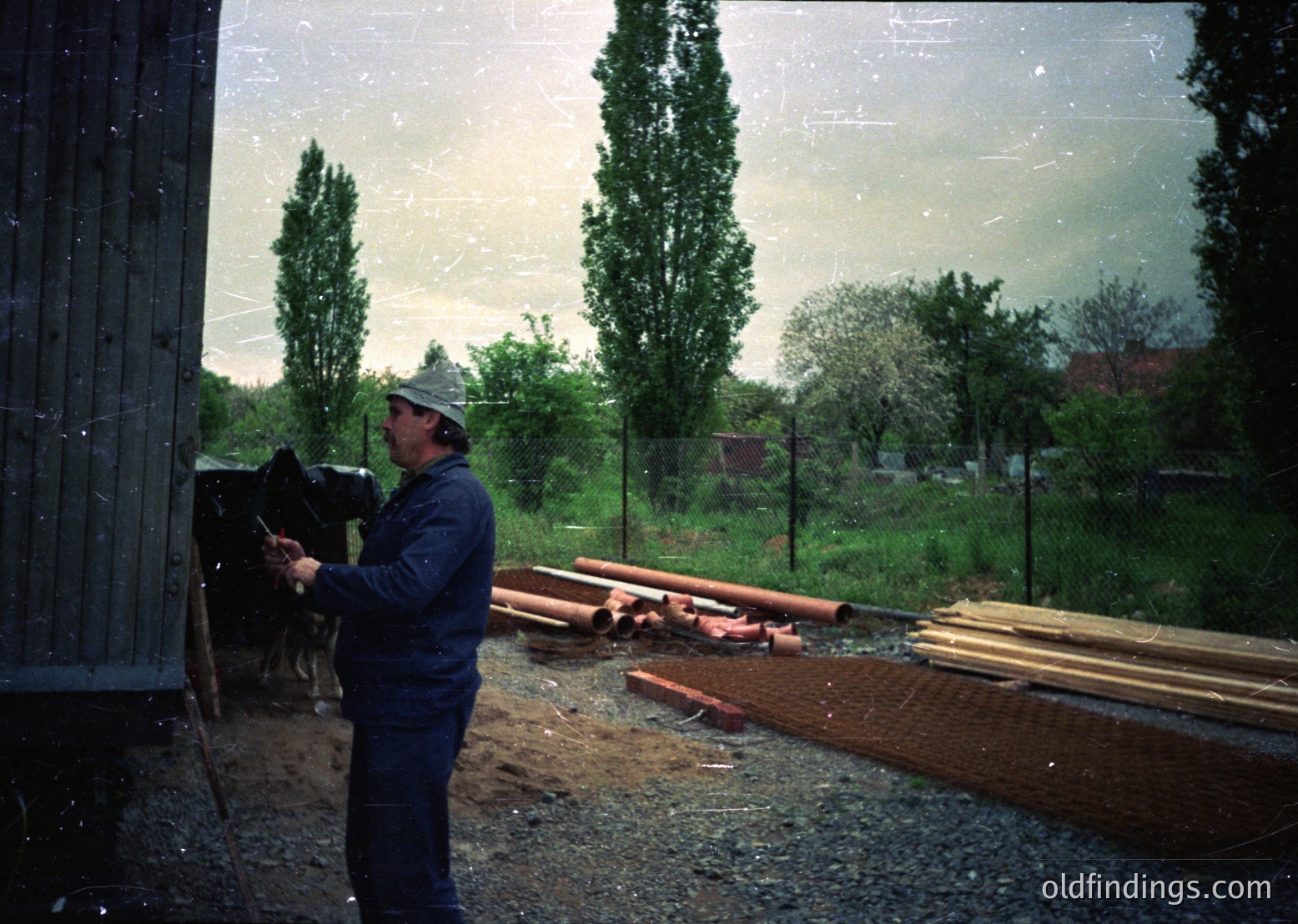 Worker in blue uniform and cap inspects rusted pipes and wooden planks near a truck, likely at a construction or maintenance site. Overcast sky and greenery suggest an outdoor setting in a temperate climate. Color tones and grain indicate a mid-20th century photograph.