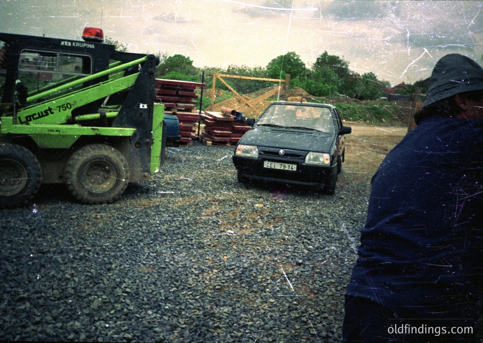 Vintage construction site with a green **Lutec 750** loader (Krupina brand) and a mid-1980s **Volkswagen Golf** parked on gravel. Overcast sky and scattered construction materials suggest mid-development. Image likely from Eastern Europe, 1980s–1990s.