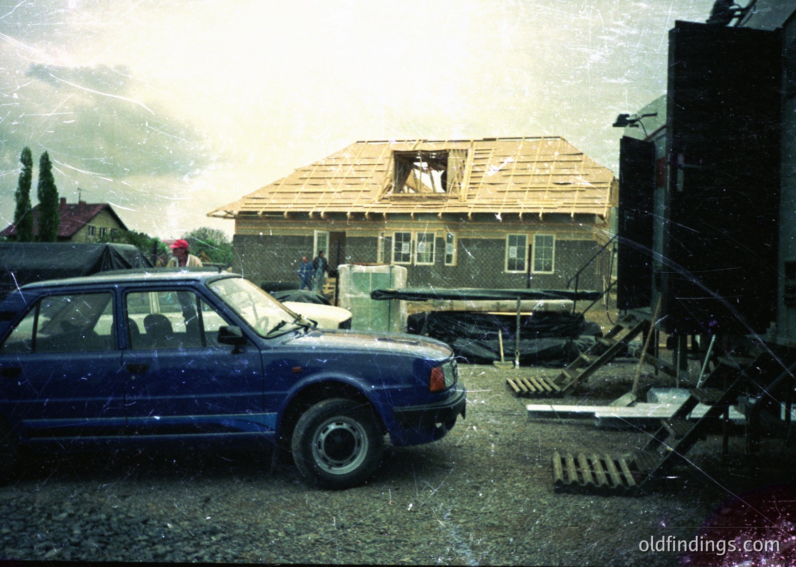 Vintage blue sedan parked beside a partially demolished single-story house under exposed roof framing. Construction debris and scaffolding indicate mid-renovation. Overcast skies suggest late 20th-century Eastern European setting.