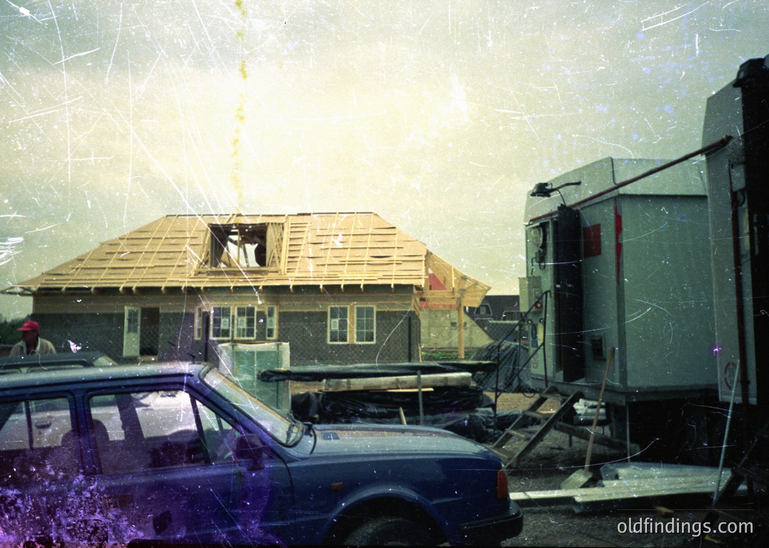 Vintage photo of a partially constructed house with exposed wooden framing and brickwork, likely mid-construction in the 1970s-1980s. A blue station wagon and a portable generator sit nearby, indicating temporary power. Heavy rain and overcast skies suggest harsh weather conditions.