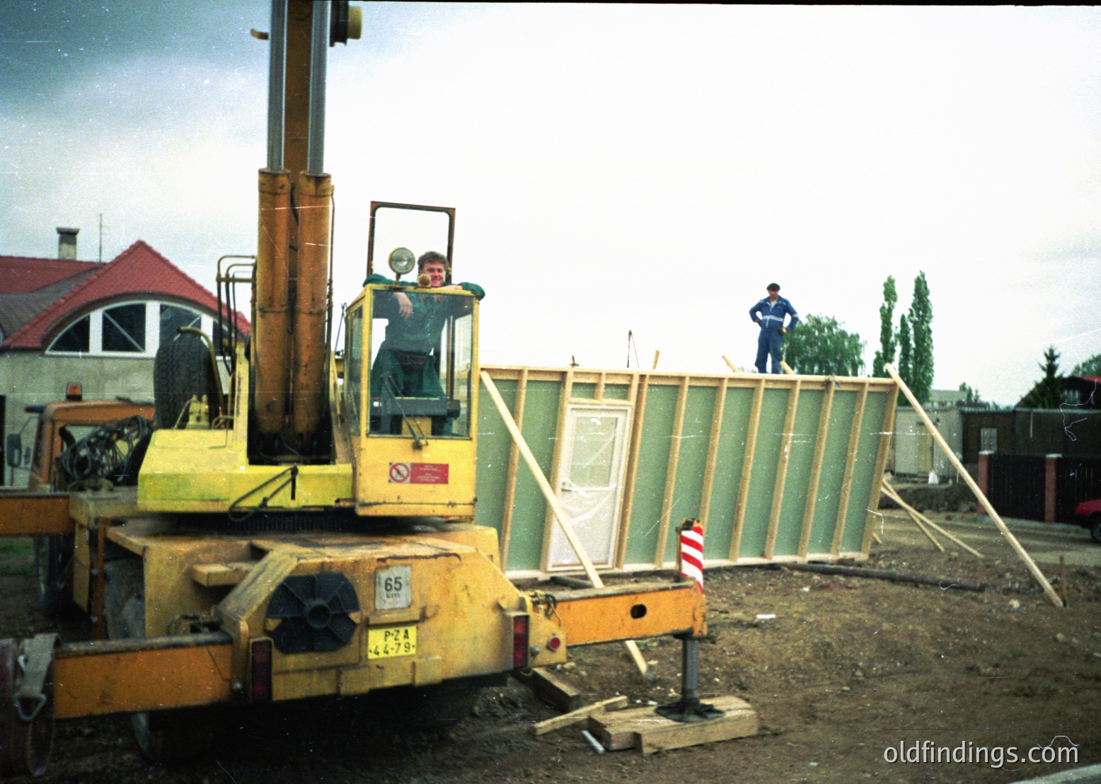 Construction site featuring a yellow hydraulic lift truck (model ) with a worker elevated in the bucket. Another worker stands on ground near a green temporary wall. Red-and-white striped barrier tape and a red-roofed building in background. Likely mid-20th century industrial or urban setting.
