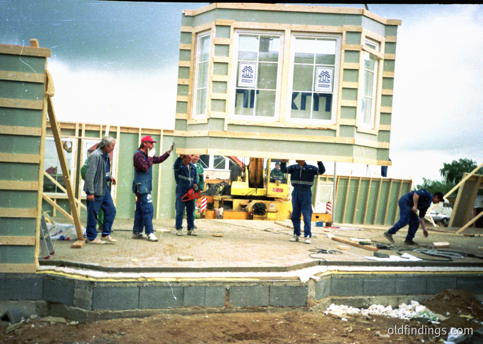 Mid-construction 2-story building with exposed wooden framing, concrete foundation, and partial brickwork. Four workers in hard hats and safety gear pose near a yellow crane. Decorative window surrounds and signage visible on upper floor. Likely 1980s-1990s industrial/commercial site.