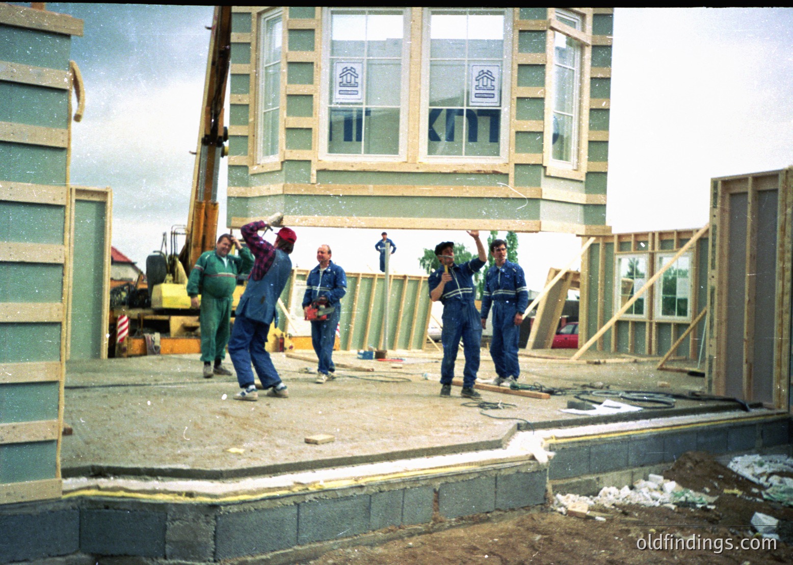 Construction crew collaboratively lifts prefab concrete beams at mid-rise building site, featuring Soviet-era architectural style. Workers wear blue overalls and hard hats; crane and scaffolding visible. Likely Eastern Bloc, 1970s-1980s.