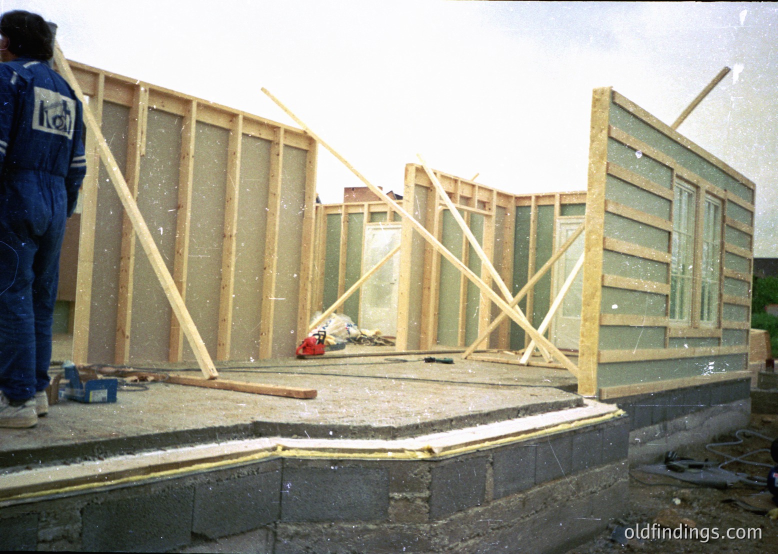 Construction frame under assembly with wooden beams and glass panels, likely mid-1980s–1990s. Worker in blue uniform (possibly industrial or utility) stands near scaffolding. Concrete foundation with exposed rebar. Urban or industrial site with partial brick wall visible.