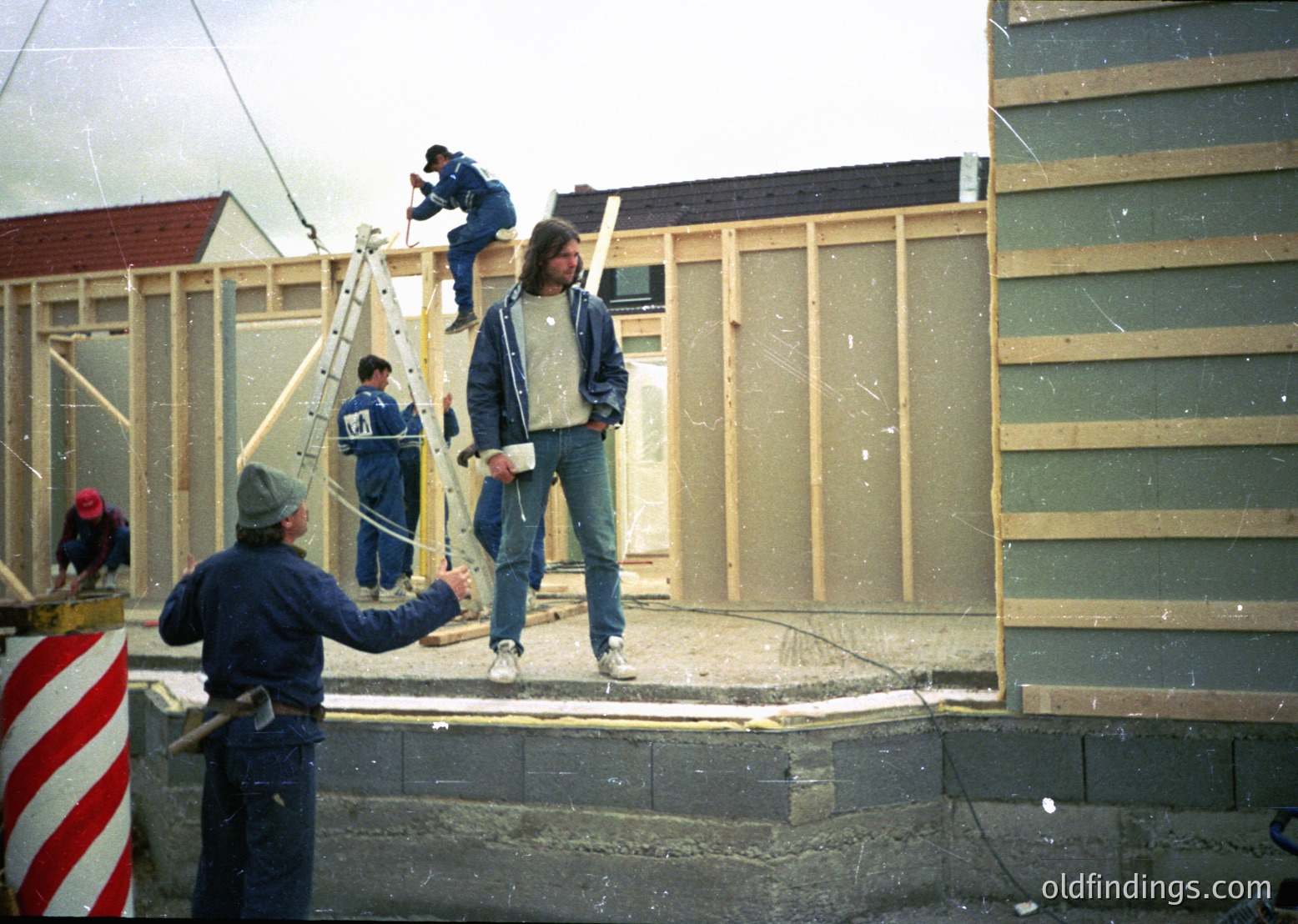 Construction crew erects prefab panel walls, 1970s-80s Eastern Bloc style. Workers in matching blue overalls and hard hats assemble modular housing units, likely for state-sponsored housing projects. Noticeable: scaffolding, concrete foundation, and industrial-era safety gear.