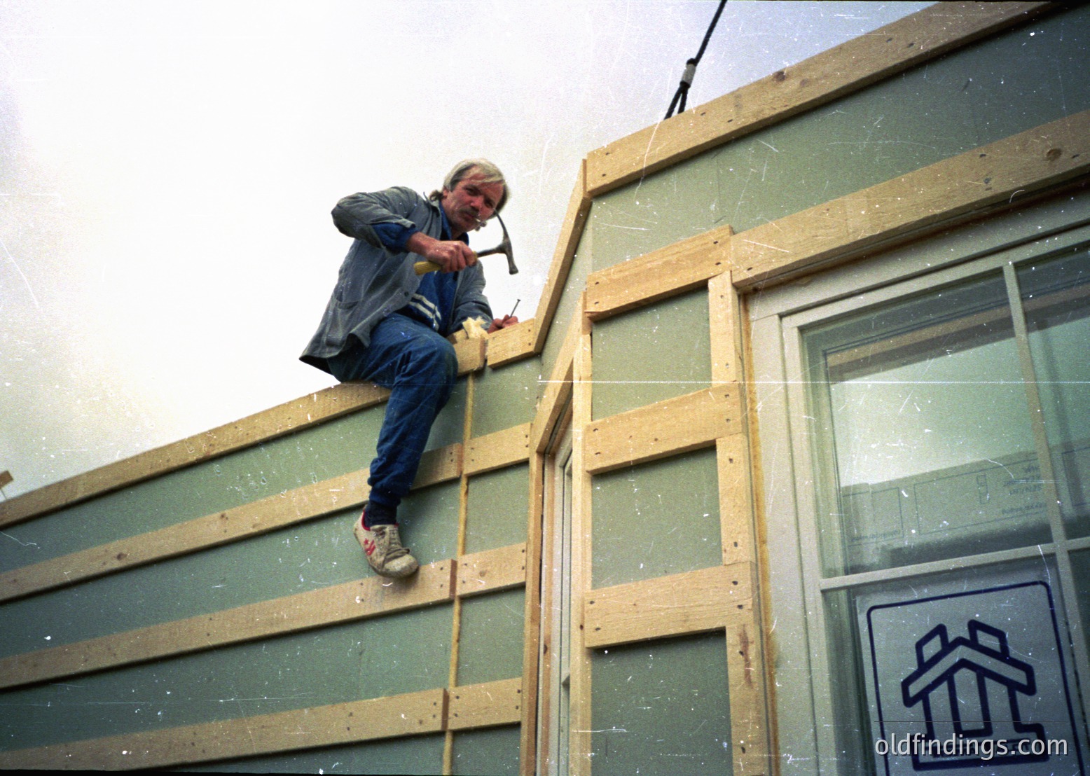 Mid-20th century construction worker securing plywood sheathing on a building frame. Overalls, tool belt, and safety-conscious stance highlight manual labor practices. Signage suggests residential development ( )