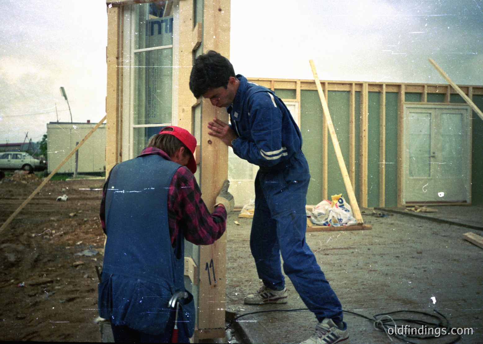 Two construction workers assemble prefab panels in a temporary site, likely 1980s–1990s Eastern Europe. Worker in red cap holds a panel while the other secures it with tools. Overcast sky and unfinished modular structures suggest rapid housing development.