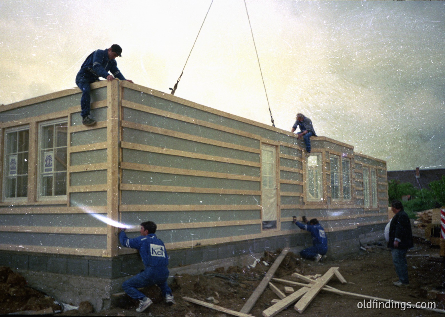 Construction crew erects prefabricated modular home, likely mid-1990s–2000s. Workers in matching blue uniforms secure large wooden panels via crane. Foundation shows poured concrete blocks; windows and doors pre-installed. Urban/suburban setting with scattered construction materials.