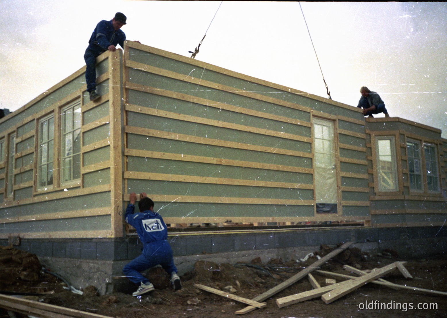 Pre-fabricated modular building construction in progress, featuring light green siding and white-framed windows. Workers in blue uniforms secure panels with ropes and planks, indicating mid-20th century industrial housing techniques. Likely Eastern Bloc-era construction ( ).