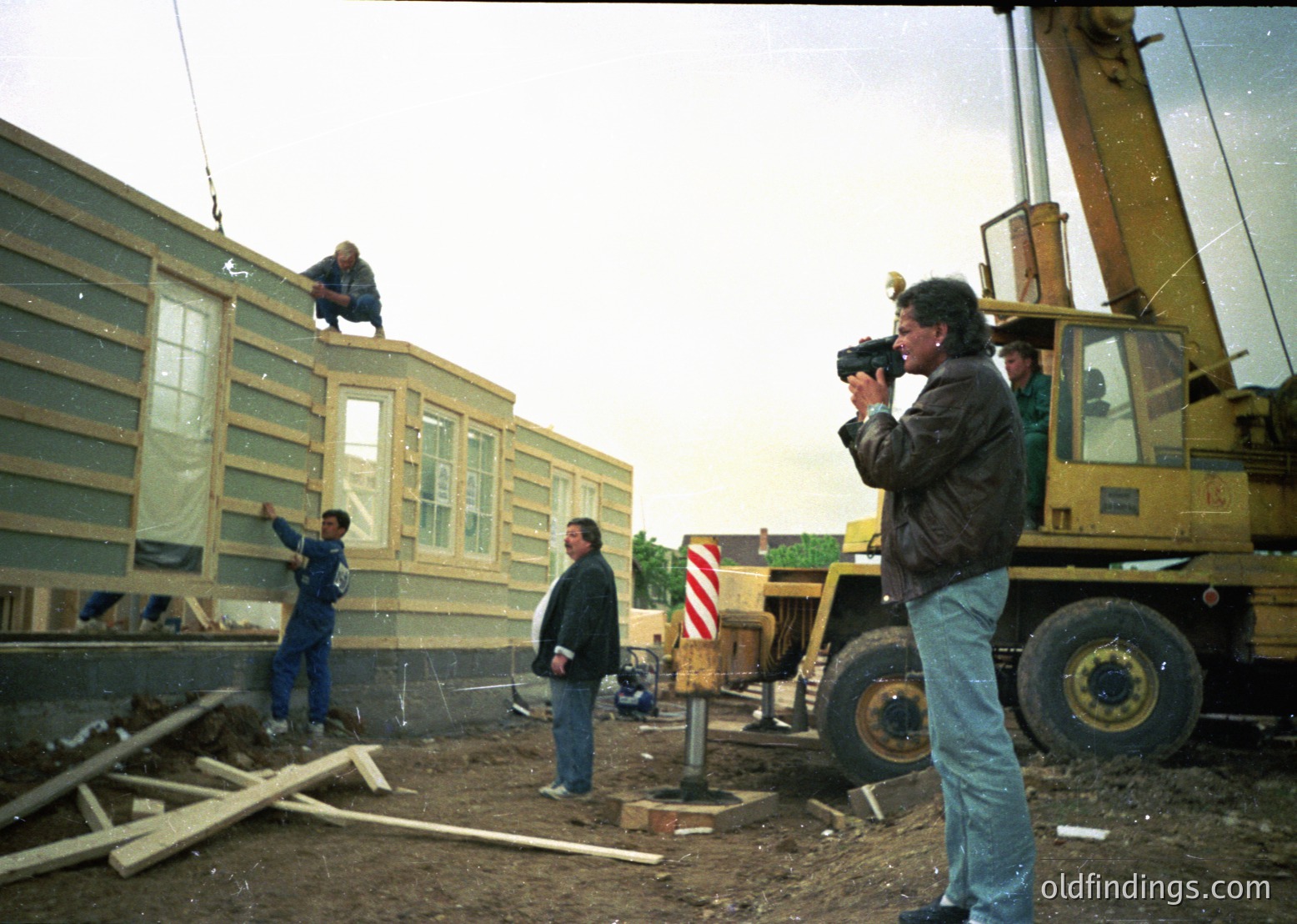 Construction site featuring prefabricated housing assembly, likely mid-1990s–2000s. Workers secure modular panels to a base, with a crane and heavy machinery visible. Photographer documents the process, capturing industrial housing development.