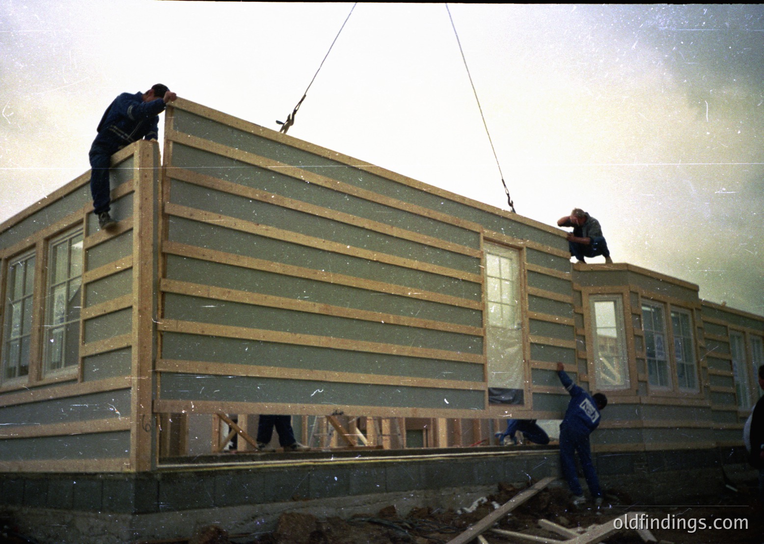Mid-20th century construction crew hoists prefab wall panels using a crane. Workers in dark uniforms secure panels to a modular building frame, likely part of post-war housing expansion. Industrial-era labor and prefabrication techniques visible.