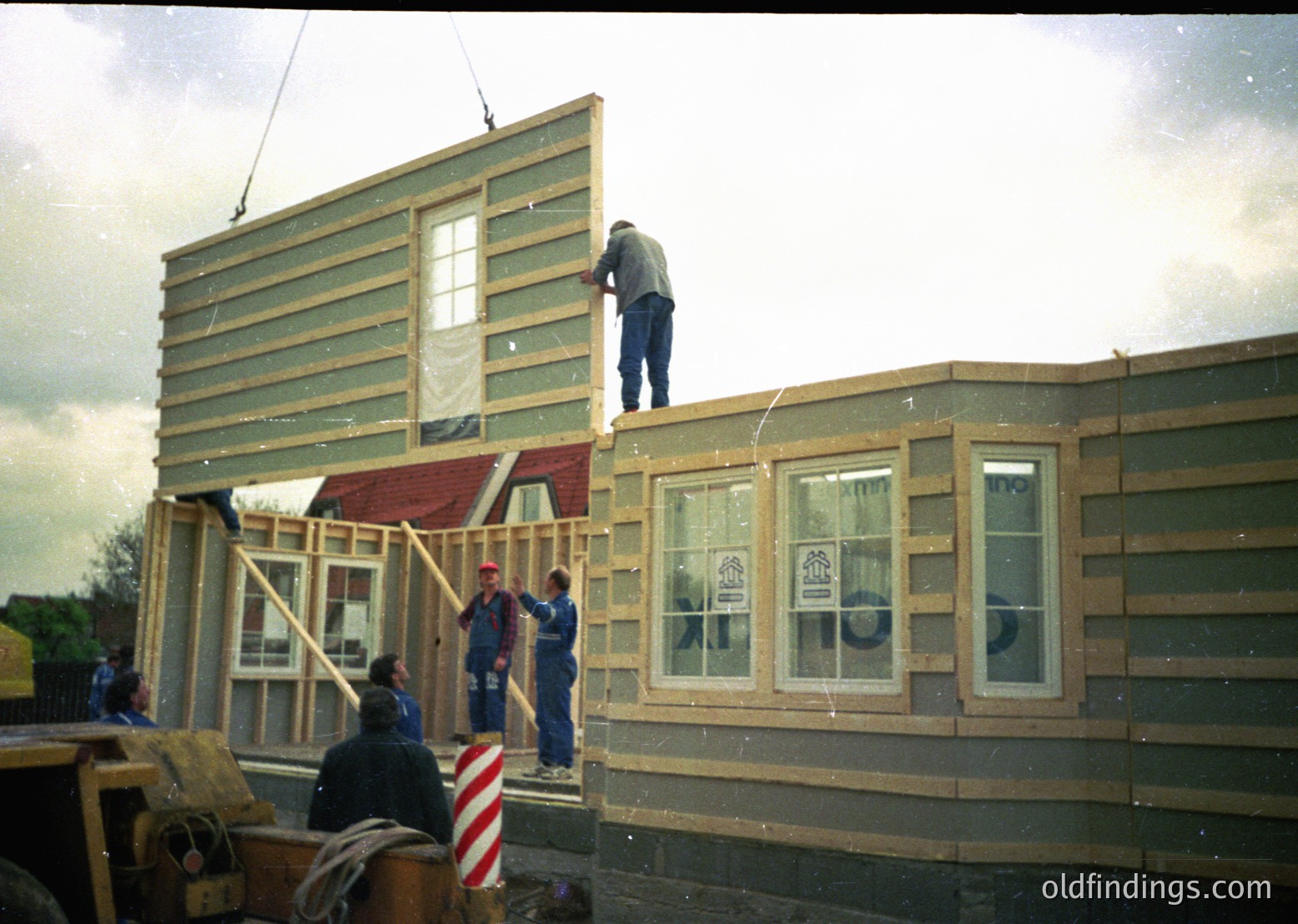 Two construction workers frame a two-story wooden house exterior, likely mid-1980s–1990s. Green siding and red roofing contrast with white-framed windows. One worker climbs the frame, while another assists below. American flag patch visible on a worker’s sleeve. Rural or suburban construction site.