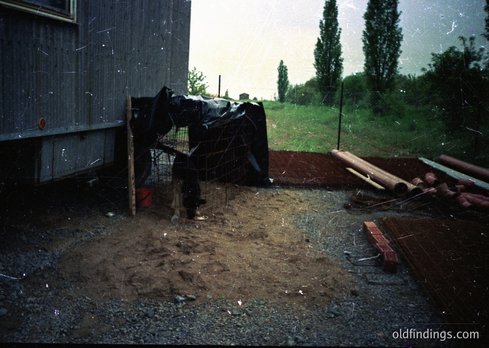 Vintage rural scene featuring a person in heavy rain gear standing near a makeshift fence and tarpaulin-covered structure. Muddy ground, scattered construction materials, and a dark blue vehicle in background suggest post-industrial or agricultural setting. Likely late 20th century.