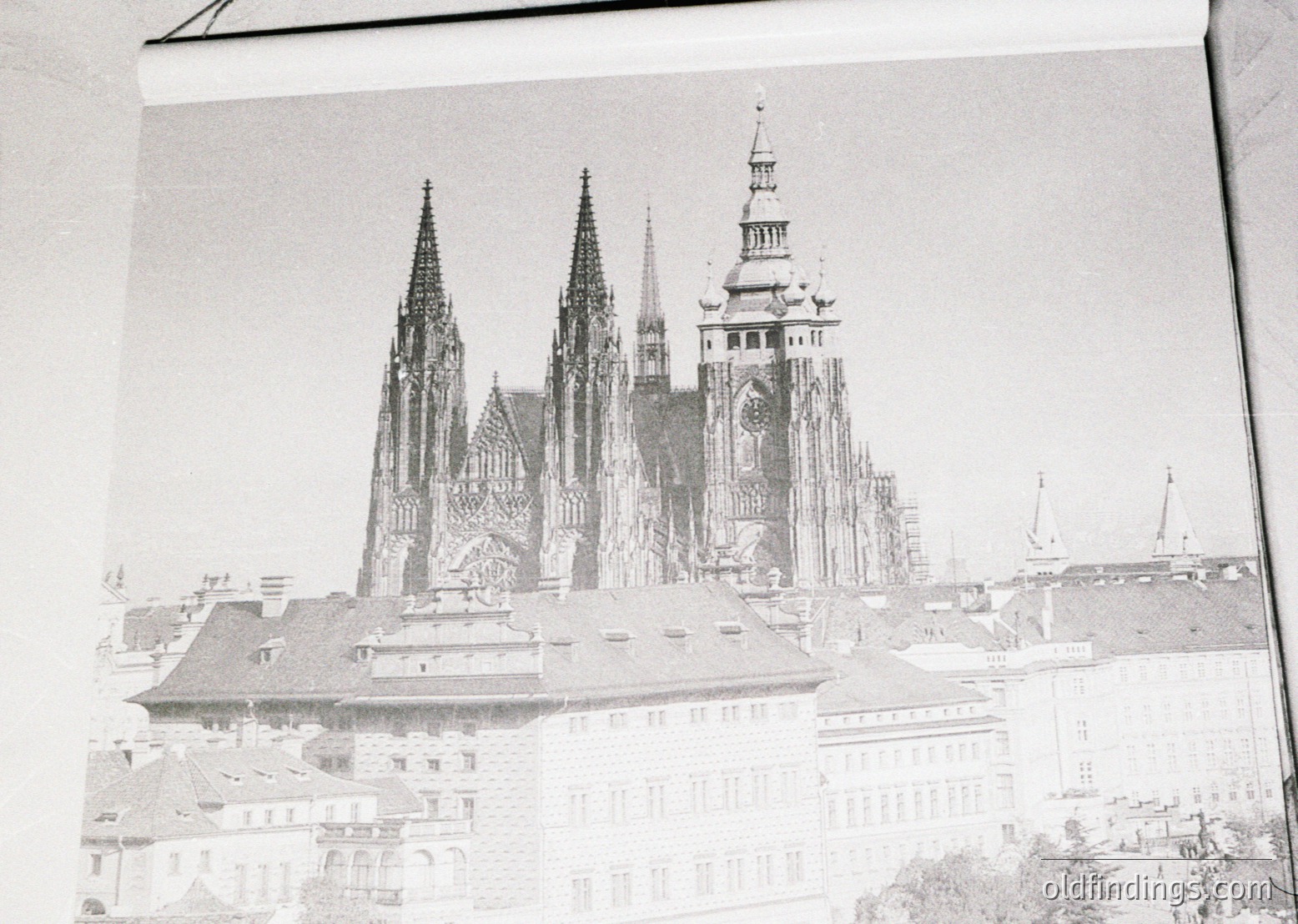 Historic Gothic cathedral with twin spires and central tower, likely **Prague Castle’s St. Vitus Cathedral**. Architectural details include pointed arches, intricate stonework, and a mix of brick and stone construction. Foreground shows low-angle perspective of adjacent buildings, suggesting urban European setting. Likely mid-20th century (1940s–1960s) based on monochrome and framing.