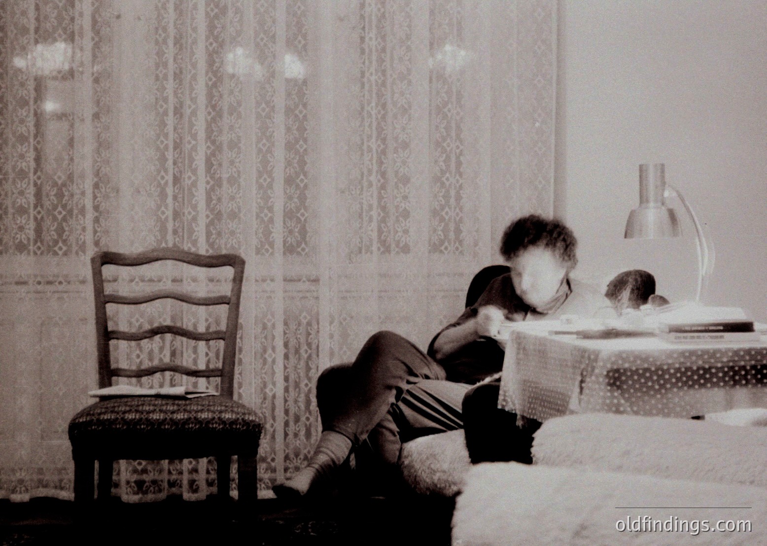 Mid-20th-century bedroom scene: child seated on patterned bedspread, engrossed in a book. Wooden chair with curved backrest and woven seat beside. Curtains with intricate lace pattern frame window. Table lamp with cylindrical shade on nightstand. Vintage monochrome aesthetic.