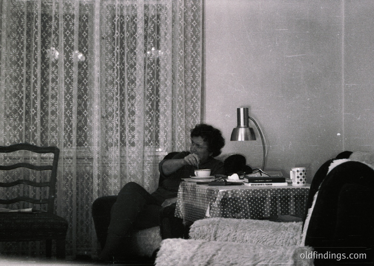 Mid-century bedroom scene featuring a person seated on a bed, drinking from a cup. Decor includes patterned curtains, a table lamp with a cylindrical shade, and a tablecloth with a polka-dot design. Folded blanket and armchair visible. Likely 1950s–1960s domestic interior.