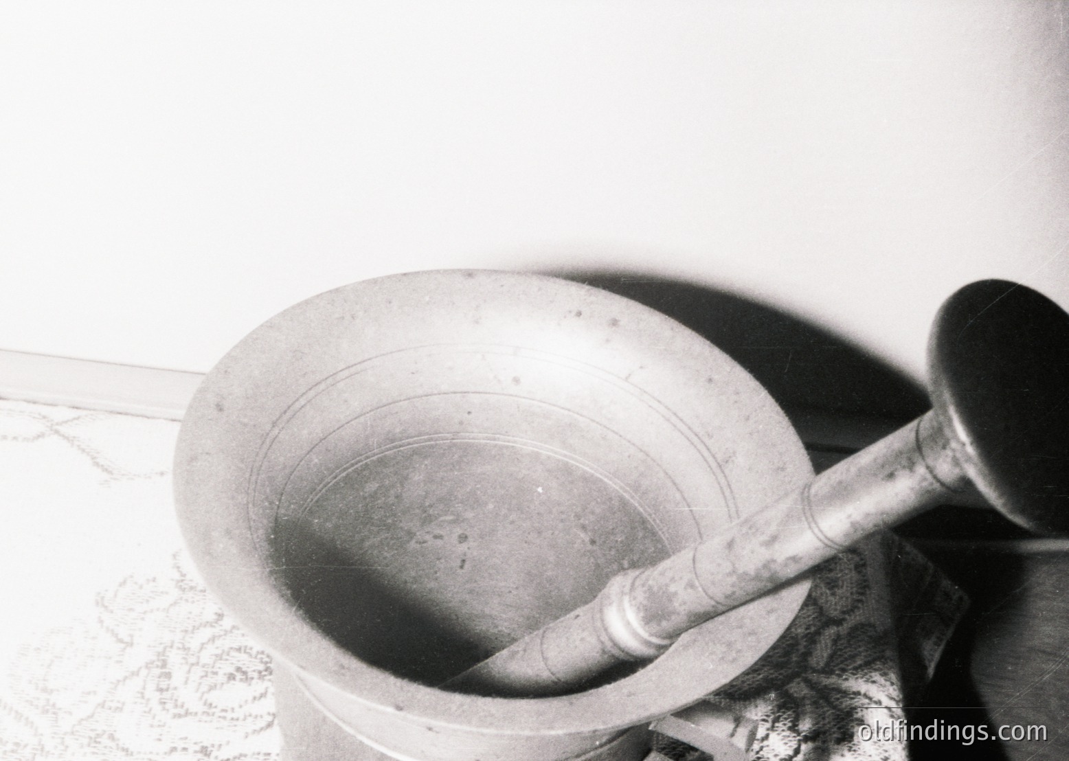Vintage mortar and pestle in use, likely for grinding herbs or spices. The wooden pestle shows wear, suggesting frequent use. Background includes a patterned cloth and blurred text, possibly a recipe or label. Ideal for historical cooking or herbalism references.
