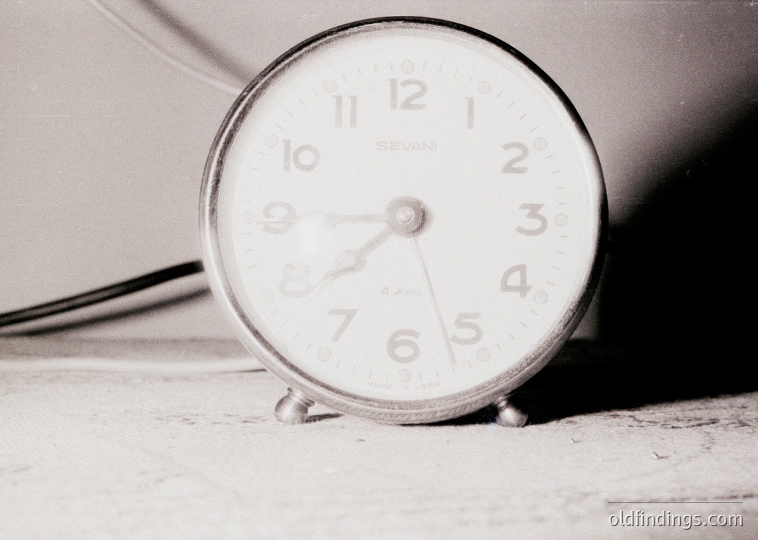 Vintage analog alarm clock with "Sevani" branding, showing 10:10. Minimalist design with three metal legs on a flat surface, black-and-white monochrome. Likely mid-20th century industrial or home decor.