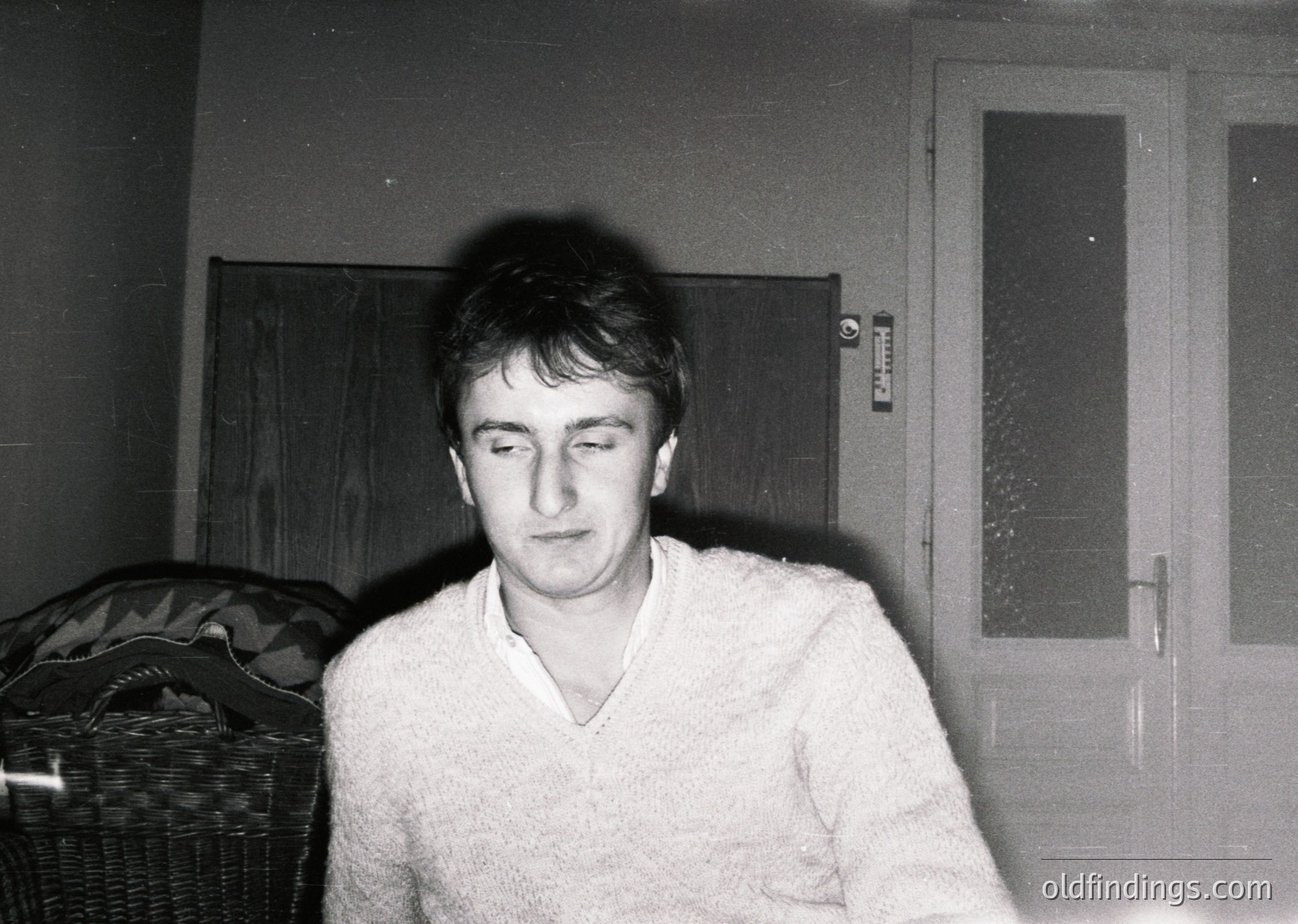 Black-and-white portrait of a man in a textured sweater, seated indoors near a wicker basket and frosted glass door. Mid-20th century interior design with functional, utilitarian elements. Likely Eastern European based on style.